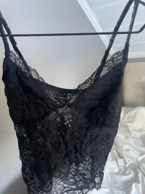 Svart spetslinne med smala axelband - Säljer ett svart spetslinne med smala axelband och v-ringad hals. Hela toppen är i genombruten spets med fina detaljer längs kanterna. Perfekt för att styla lager på lager eller som statement-piece.