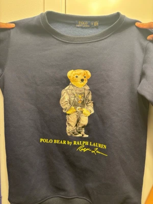 Blå sweatshirt Polo Bear Ralph Lauren - Mörkblå sweatshirt från Polo Ralph Lauren med ikoniska Polo Bear-trycket framtill. Tröjan har rund halsringning och ribbade muddar vid ärmslut och nederkant. Tillverkad i mjukt bomullsmaterial och har en avslappnad passform. Perfekt för en casual och trendig look. OBS! Priset går alltid att få ner 