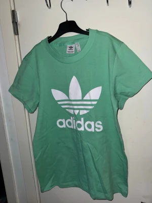 Grön Adidas t-shirt med logga - Snygg grön t-shirt från Adidas med den klassiska vita trefoil-loggan tryckt framtill. T-shirten har rund halsringning och korta ärmar. Tillverkad i mjuk bomull som känns skön mot huden. Perfekt för en avslappnad och sportig stil.