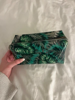 Grön necessär med palmbladsmönster - Snygg necessär med grönt palmbladsmönster på grå botten och dragkedja i guld. Perfekt för att förvara smink, småprylar eller andra viktiga saker. Ganska använd och lite smutsig på insidan 