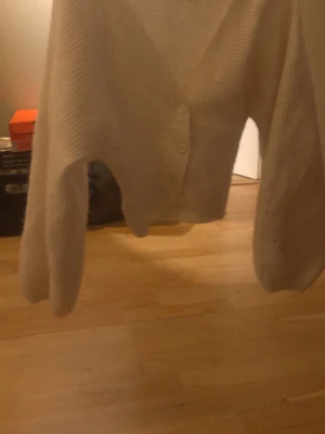 Beige stickad kofta med v-ringning - Mysig beige stickad kofta med v-ringning och två knappar framtill. Koftan har långärmade ärmar och en avslappnad passform, perfekt att slänga på sig över en t-shirt eller topp. Materialet är mjukt och ger en skön känsla.