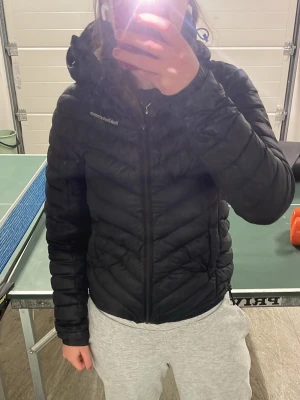 Svart dunjacka från Peak Performance - Säljer en svart dunjacka från Peak Performance med huva och dragkedja framtill. Jackan har ett stilrent, quiltat mönster och logga på bröstet och axeln. Perfekt för vinterdagar och är riktigt skön att bära. Passar dig som vill ha både värme och en snygg look.