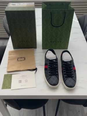 Gucci GG Supreme sneakers svart/röd - Snygga Gucci sneakers i svart GG Supreme canvas med klassiska röda och blå ränder på sidan. Skorna har vita sulor, svarta skosnören och vita innersulor med Gucci-logga. Perfekta för dig som vill ha en exklusiv och trendig look.