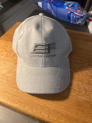 Grå EF Limited Edition keps - Snygg grå keps från EF med broderad logga och texten 'Limited Edition' framtill. Klassisk böjd skärm och justerbar baksida. Tillverkad i ett mjukt tygmaterial som ger en clean och stilren look.