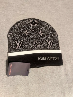 Svart Luis Vuitton mössa - Snygg svart och vit mössa från Luis Vuitton med klassiskt monogrammönster och broderad logga på uppviket. Tillverkad i mjukt stickat material som känns lyxigt och trendigt. Perfekt accessoar för dig som vill sticka ut med exklusiv stil.