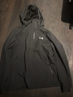 Svart vindjacka från Under Armour - Snygg svart vindjacka från Under Armour med huva och dragkedja framtill. Jackan har två sidofickor med dragkedja och logga på bröstet. Tillverkad i ett lätt och slitstarkt material som står emot vind. Perfekt för dig som vill ha en sportig och stilren look.