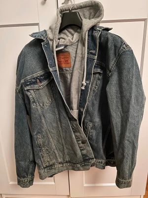 Levi's x Vetements blå jeansjacka med hoodie - Säljer en oversized blå jeansjacka från Levi's x Vetements  med grå sweatshirt-hoodie insydd. Jackan har två bröstfickor med lock, sidofickor och knappstängning framtill. Oversized passform och cool streetwear-vibe. Perfekt för lager-på-lager-stil.