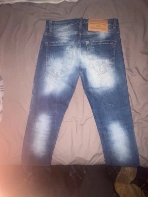 Blå slitna dsquared jeans med hål och stänk - Säljer ett par blå jeans från DSQUARED2 med slitningar, stora hål och vita färgstänk på framsidan. Jeansen har en tvättad look med ljusa partier och två bakfickor. Perfekta för dig som gillar en edgy och trendig stil.