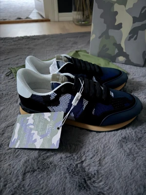 Valentino Garavani Rockrunner sneakers - Snygga Valentino Garavani Rockrunner sneakers i blått, svart och vitt med camo-mönster. Skorna har mesh och mocka-paneler, svarta snören och beige sula. Ikonisk nitar-detalj på hälen och vit hälkappa. Perfekta för dig som vill sticka ut med exklusiva sneakers.
