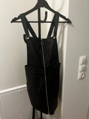 Svart kjol-jumpsuit med dragkedja - Snygg svart kjol-jumpsuit med breda axelband och justerbara spännen. Framtill finns en lång silverfärgad dragkedja som detalj. Modellen har fickor på sidorna och är tillverkad i ett glansigt tyg som ger en cool look. Perfekt för dig som vill ha en edgy stil.