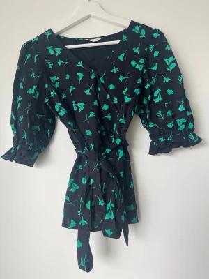 Svart blommig omlottklänning med knyt - Snygg svart kortklänning med grönt blommönster och omlottmodell. Klänningen har trekvartsärm med volangkant och knyt i midjan för en smickrande passform. Perfekt för dig som gillar trendiga detaljer och en feminin look.