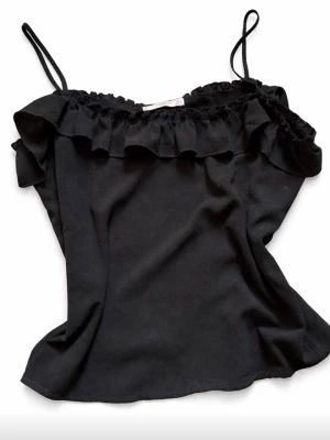 Svart volangtop med smala band - Snygg svart topp med tunna axelband och volangdetalj längs urringningen. Stolek S från Mango. 
