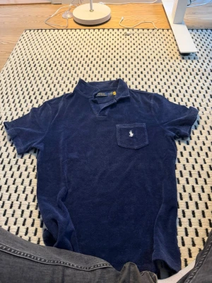 Blå frottéskjorta från Polo Ralph Lauren - Mörkblå kortärmad skjorta i mjuk frotté från Polo Ralph Lauren. Skjortan har klassisk krage, bröstficka med broderad logga och avslappnad passform. Perfekt för en chill och stilren look.