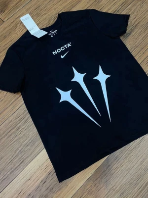 Svart NOCTA x Nike t-shirt - Snygg svart t-shirt från Nike och NOCTA med stora vita grafiska tryck framtill och logga på bröstet. Klassisk passform och rund halsringning. Perfekt för dig som gillar streetwear och vill sticka ut.