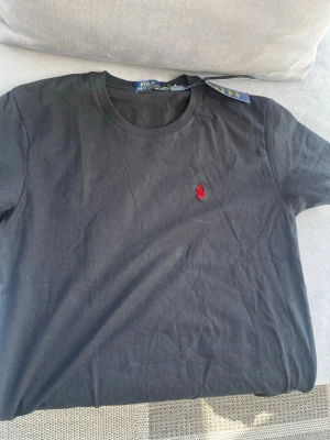 Svart t-shirt från Polo Ralph Lauren - Klassisk svart t-shirt från Polo Ralph Lauren med rund hals och ikonisk röd logga broderad på bröstet. Tillverkad i mjuk bomull för en skön och avslappnad passform. Perfekt basplagg som funkar till allt.