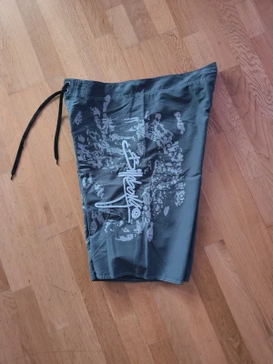 Gråa shorts från Billabong - Säljer ett par gråa shorts från Billabong med vit grafisk logga och mönster på sidan. Shortsen har snörning i midjan och en ficka med lock på sidan. Snygga till allt