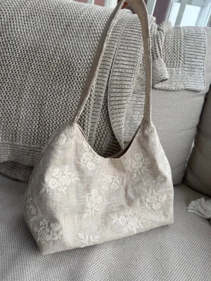 Beige axelremsväska med broderi - Säljer en beige handväska i linneliknande material med vackra vita blommiga broderier. Väskan har en mjuk, avlång form och bred axelrem som gör den enkel att bära. Perfekt för dig som gillar en stilren och naturlig look.