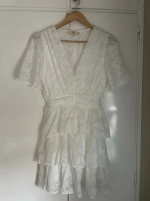 Vit kortklänning från Chelsea - Supersöt vit kortklänning från Chelsea med v-ringning, volangdetaljer och korta ärmar. Klänningen har ett subtilt blommigt mönster och omlottkänsla i midjan. Perfekt för sommarens alla tillfällen!