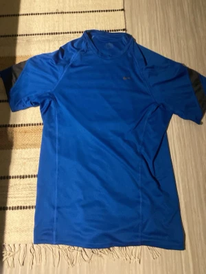 Blå Nike träningströja - Snygg blå träningströja från Nike med korta ärmar och svart detalj på ärmarna. Tillverkad i ett lätt och snabbtorkande funktionsmaterial som passar perfekt till träning och sport. Klassisk Nike Swoosh-logga på bröstet.