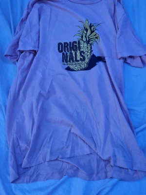 Ljuslila t-shirt Jack & Jones Originals - Säljer en ljuslila t-shirt från Jack & Jones Originals med coolt ananastryck och texten 'Originals' på bröstet. T-shirten har korta ärmar och är gjord i mjuk bomull, perfekt för en avslappnad stil.