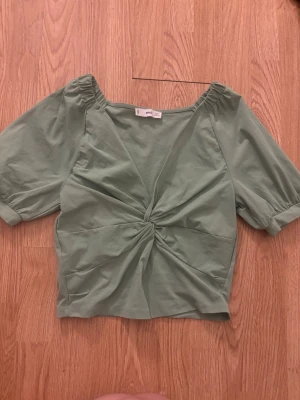 Grön v-ringad topp med twist från MNG - Snygg ljusgrön topp från MNG med v-ringning och twistad detalj framtill. Toppen har korta puffärmar med resår vid axlarna och är tillverkad i mjuk bomull. Perfekt för dig som vill ha en trendig och bekväm look.