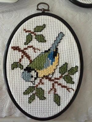 Broderad tavla med blåmes - Säljer en oval broderad tavla med motiv av en blåmes på en gren med gröna blad. Tavlan har svart ram och broderiet är gjort på vit aidaväv med garn i blått, gult, grönt, brunt och svart. Perfekt som unik väggdekoration.