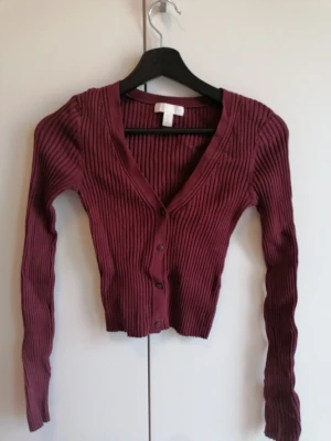 Vinröd ribbad tröja - Vinröd ribbstickad tröja med djup V-ringning och knäppning framtill. Långärmad, kort och figurnära modell som passar perfekt till jeans eller kjol. Mjuk viskos och stretchig i materialet för en bekväm känsla.