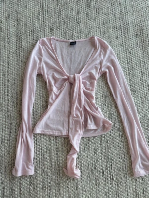 Ljusrosa knytblus från Gina Tricot - Supersöt ljusrosa blus från Gina Tricot med lång ärm och v-ringning. Toppen har en snygg knytning framtill som ger en feminin touch. Tyget är luftigt som är perfekt till sommaren. 