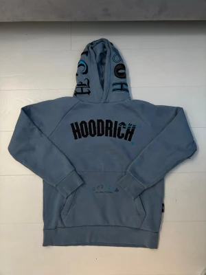 Blå hoodie från Hoodrich med tryck. Pris kan diskuteras!!!! - Snygg blå hoodie från Hoodrich med stor svart logga på bröstet och coola tryck på huvan. Mjuk bomullskänsla och känguruficka framtill. Baksidan har texttryck i svart. Perfekt för en streetwear-look.
