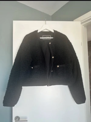 Svart teddyjacka med silverknappar - Helt ny Mysig svart teddyjacka från med rund halsringning och raka ärmar. Jackan har dekorativa knappar framtill och två fickor med knappdetaljer. Perfekt för dig som vill ha en trendig och bekväm look i höst och vinter.