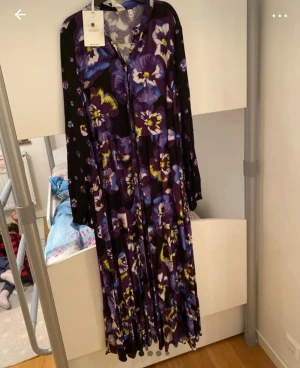 Långklänning med lila blommor från Y.A.S - Långklänning från Y.A.S med stort blommigt mönster i lila, blått och gult på svart botten. Klänningen har v-ringning, långa ärmar och ett luftigt fall. Perfekt för dig som gillar färgstarka och unika plagg. Nu pris:1049kr