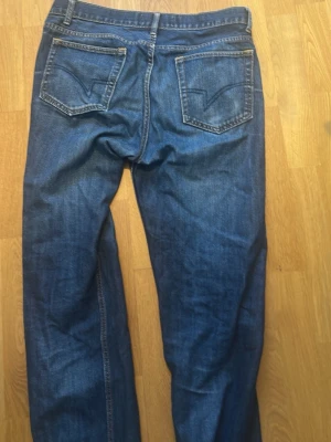 Blå raka jeans Dressman - Säljer ett par klassiska blå jeans med raka ben och snygga broderade bakfickor. Jeansen har normal midja och är tillverkade i denim med tydliga sömmar. Perfekta för en avslappnad och stilren look.