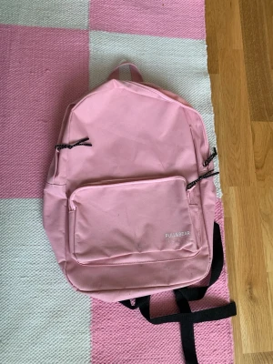 Rosa ryggsäck från Pull&Bear - Snygg ljusrosa ryggsäck från Pull&Bear med svartvita dragkedjor och justerbara svarta axelband. Rymligt huvudfack och en stor ytterficka framtill. Perfekt för skolan eller stan, tillverkad i slitstark textil. Fläckar förekommer, se på bild.