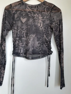 Mesh topp med änglamotiv och snörning - Snygg mesh topp i mörkgrått med detaljerat änglamotiv och hjärtan. Toppen har lång ärm och cool snörning i sidorna för extra edge. Materialet är tunt och stretchigt, perfekt för lager på lager. Ger en alternativ vibe till din outfit.