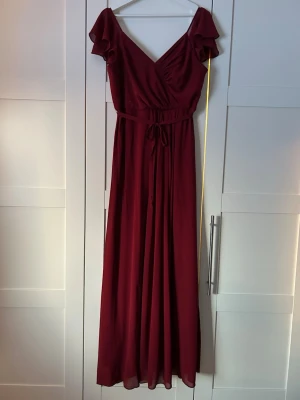 Vinröd långklänning med volangärm - Elegant vinröd långklänning med omlott upptill, v-ringning och volangärmar. Klänningen har ett knytband i midjan som framhäver siluetten och ett luftigt, svepande tyg. Perfekt för speciella tillfällen. Jag är 160cm och hade på mig 9cm klackar på bilden. Använd endast en gång, under studentbalen. 
