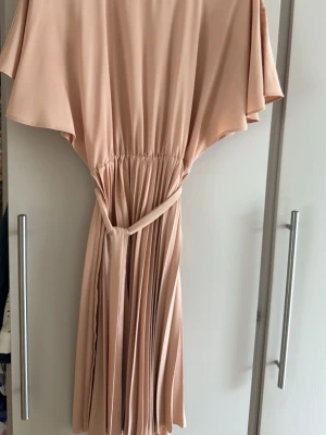 Ljusbeige omlottklänning med volang - Säljer en ljusbeige klänning med omlottknytning i midjan och vackra volangärmar. Kjolen är plisserad och klänningen har en mjuk, flowy känsla. Perfekt för dig som gillar en feminin och stilren look.