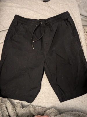 Svarta shorts med snörning stl 164 - Svarta shorts med elastisk midja och snörning framtill. Två sidofickor och två bakfickor med lock. Perfekta för sommaren och har en avslappnad passform. Materialet känns som bomull och är mjukt mot huden.