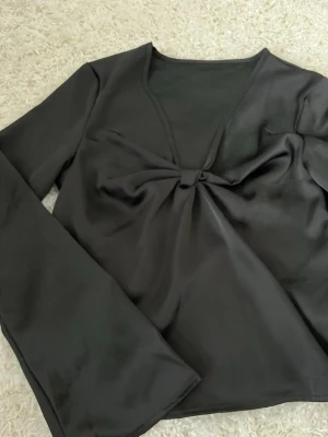 Svart långärmad topp med rosett - Svart långärmad topp med v-ringning och snygg rosettdetalj framtill. Tunn och mjuk känsla, perfekt för att lyfta en enkel outfit. Sitter snyggt och har en stilren look.