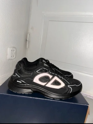 Svarta Dior sneakers med CD-logga - Snygga svarta sneakers från Dior med stor vit CD-logga på sidan. Skorna har en chunky sula, svarta snören och detaljer i vitt och grått. Ovandelen är i mesh och syntetmaterial för en modern look.