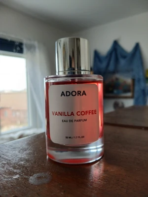 Adora Vanilla Coffee Eau de Parfum - En parfymflaska från Adora med doften Vanilla Coffee. Flaskan är cylindrisk i genomskinligt glas med en metallisk silverfärgad kork. Vätskan inuti är röd och etiketten är vit med röd text. Innehåller 50 ml Eau de Parfum.❤️