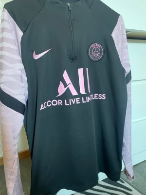 PSG träningströja svart/lila Nike - Snygg långärmad träningströja från Paris Saint-Germain i svart med lila detaljer och logga. Tröjan har halv dragkedja, vågmönstrade ärmar och tryckt Nike-logga samt sponsortryck på bröstet. Perfekt för fotbollsträning eller sportiga dagar.