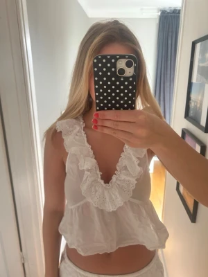 Vit volangblus med broderad spets - Söt vit blus med djup v-ringning och volangkant i broderad spets. Modellen är ärmlös och har en luftig, croppad passform som ger en romantisk känsla. Perfekt för varma dagar och somriga outfits.