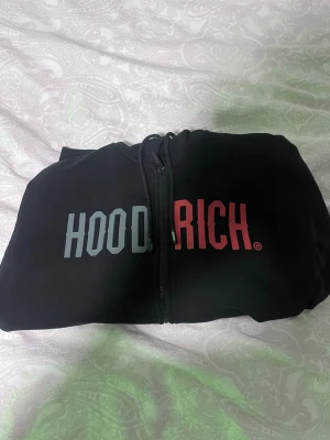 Hoodrich Hoodie - Säljer en svart zip hoodie från Hoodrich med stor logga i vitt och rött över bröstet. Hoodien har dragkedja framtill, fickor och justerbar huva med snören. Perfekt för en chill och street-inspirerad look. Materialet känns mjukt och bekvämt. Knappt använd, säljer för att jag aldrig använder den. Lite större i storleken.