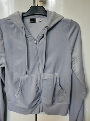 Ljusblå zip hoodie från Gina Tricot - Ljusblå hoodie i mjukt velourmaterial från Gina Tricot. Dragkedja framtill, två stora fickor och justerbar huva med snören. Croppad modell med ribbade muddar vid ärmslut och nederkant. Perfekt för en chill och avslappnad stil. Svårt att få till rätt färg men den är ljusblå.