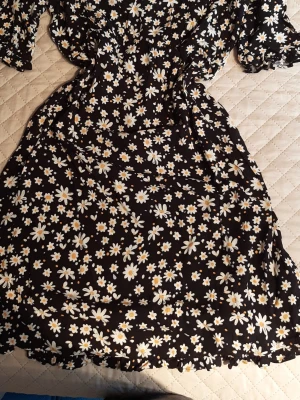 Svart blommig kortklänning - Supersöt svart kortklänning med vita och gula blommor. Klänningen har en smickrande passform med markerad midja och korta ärmar. Perfekt för varma dagar och har en lekfull vibe med sitt blommiga mönster.