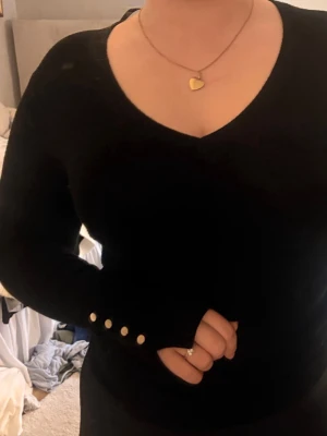 Svart långarmad tröja - Svart långärmad tröja med v-ringning och guldfärgade knappar vid ärmarna. Tröjan har en slim fit och är perfekt för lager-på-lager. Mjuk och skön känsla, passar till både jeans och kjol. 😝