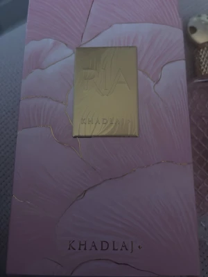 Khadlaj RIA parfym rosa och guld - Exklusiv parfym från Khadlaj i en snygg rosa förpackning med guldiga detaljer och blommigt mönster. Lådan har en lyxig känsla med en guldig platta framtill där det står RIA. Perfekt för dig som gillar eleganta och unika dofter.