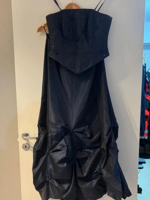Två delars klänning i marinblå - Snygg svart klänning med inga axelband och unik draperad kjol. Korsetten är dekorerat med glittriga rosetter och små tygblommor. Klänningen har en markerad midja och ballongliknande kjol för en cool siluett. Perfekt för dig som vill sticka ut.