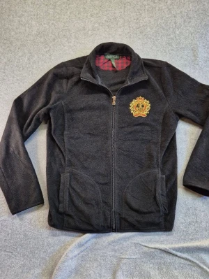 Svart fleecejacka från Ralph Lauren - En Fleece jacka från Lauren by Ralph Lauren! Snyggt brodyrmärke på bröst. Har märke i tyget över mage se bild. Se bröst och längdmått på sista bilderna så du vet den passar dig perfekt! Har även med ärmlängd och då är det mätt från övre axelsöm ned till ärmsmudd