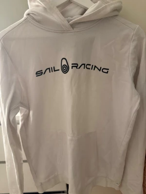 Vit hoodie från Sail Racing - Vit hoodie från Sail Racing med svart logga tryckt på bröstet. Den är använd 1 gång och är i storlek 170 vilket motsvarar runt storlek S! Den är köpt för 900kr på sail racing hemsida .Klassisk modell med huva och känguruficka framtill. Mjuk bomullskänsla och ribbade muddar vid ärmslut och nederkant. Perfekt för en avslappnad och sportig stil.
